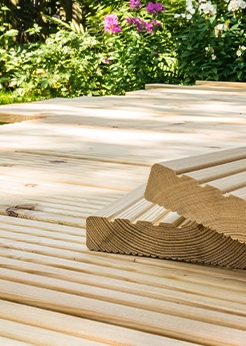 Terrassenbau mit hellen Lärchenholzdielen, gestapelte Riffelbretter im Vordergrund, umgeben von grünem Garten.