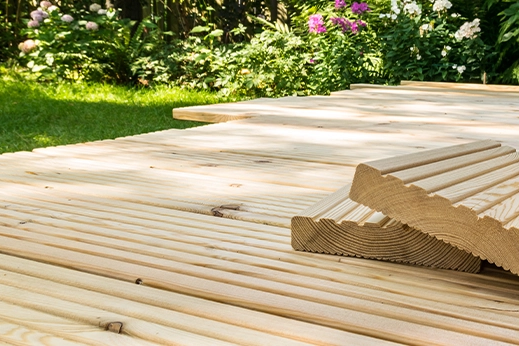 Terrassenbau mit hellen Lärchenholzdielen, gestapelte Riffelbretter im Vordergrund, umgeben von grünem Garten.