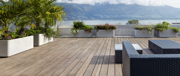 Große Terrasse mit Bambus-Holzdielen, modernen Rattanmöbeln, Pflanzen in Betonkübeln und Blick auf einen See mit Bergen.
