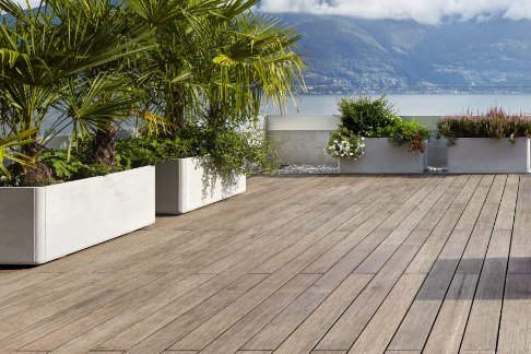 Große Terrasse mit Bambus-Holzdielen, modernen Rattanmöbeln, Pflanzen in Betonkübeln und Blick auf einen See mit Bergen.