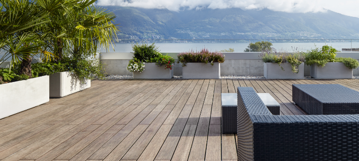 Große Terrasse mit Bambus-Holzdielen, modernen Rattanmöbeln, Pflanzen in Betonkübeln und Blick auf einen See mit Bergen.