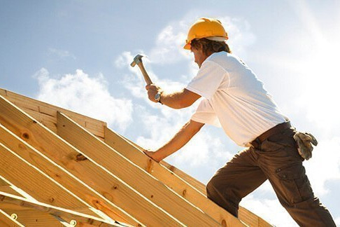 Dachdecker mit gelbem Schutzhelm beim Befestigen von Konstruktionsvollholz (KVH) auf einem Dachstuhl unter strahlendem Himmel.